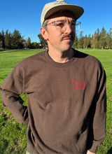 Load image into Gallery viewer, Man wearing a brown sweatshirt with a logo, standing in a grassy field with trees and a clear blue sky.